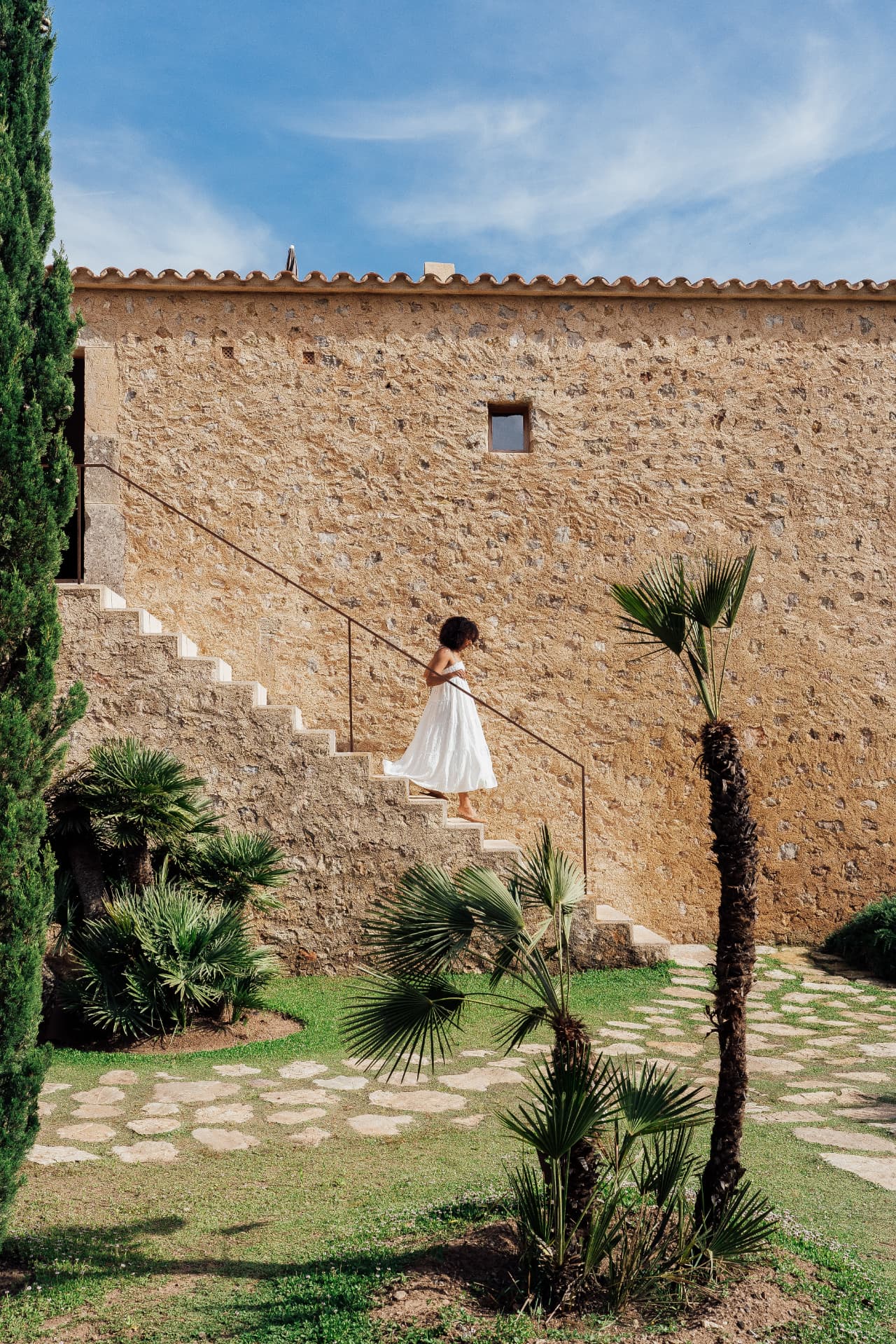 A woman in a white dress walking up stone stairs at a sunlit Mallorcan retreat